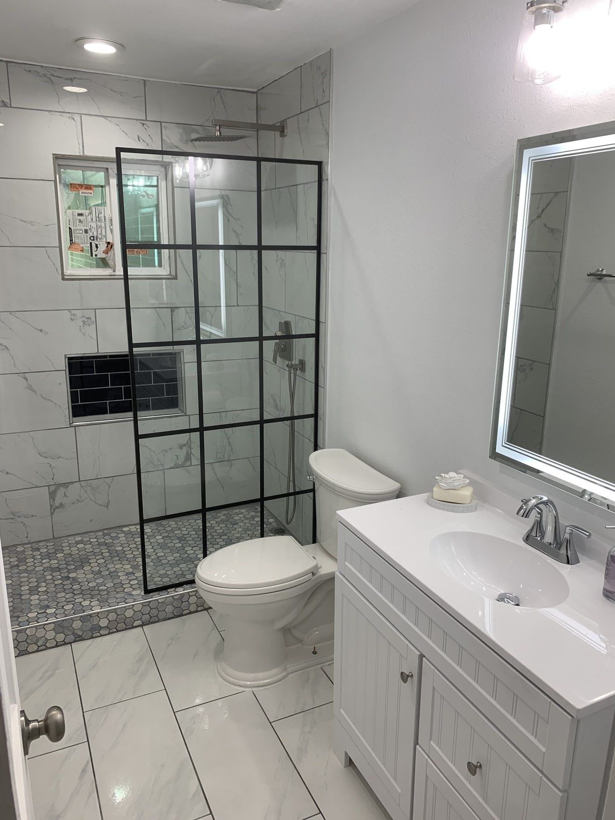 Same bathroom after the remodel, with a black grid glass shower, white vanity and lit mirror
