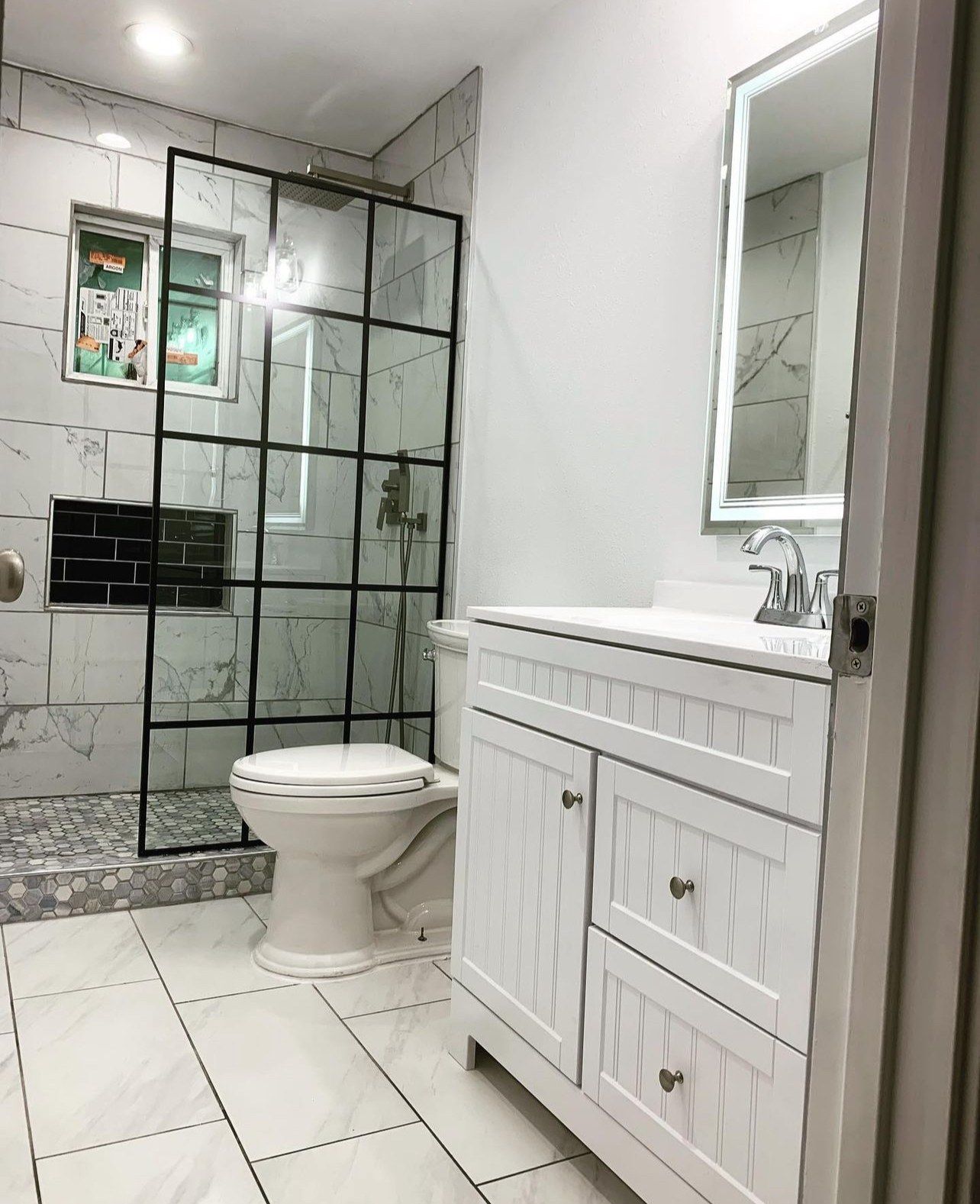 Finished bathroom with a black grid glass shower, white shaker vanity and LED mirror